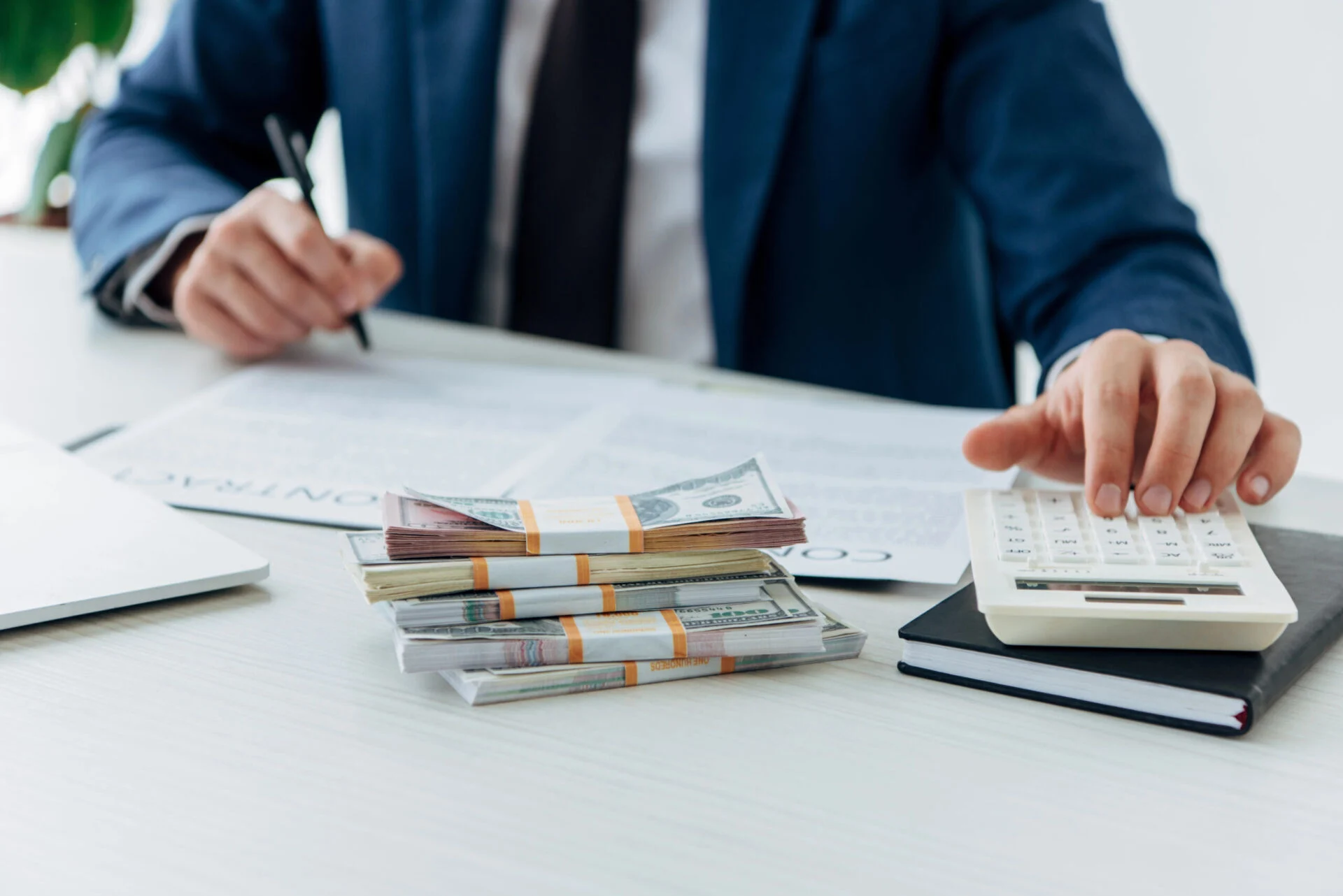 Person in a suite using a calculator while signing documents in front of a pile of money